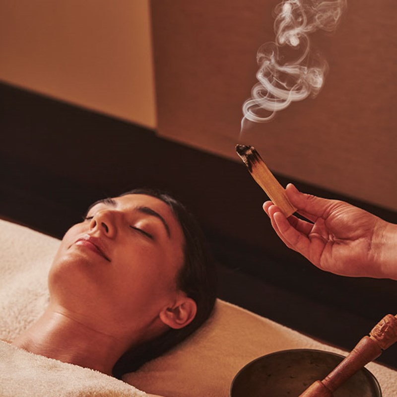 Wellness Ritual Relaxed woman on a massage table with a therapist holding smoking palo santo over her, evoking a calming spa ritual.