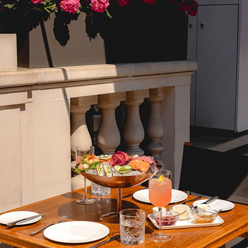 Alfresco table with seafood platter, glassware, and pink drink, set against stone balustrades and blooming pink flowers above.