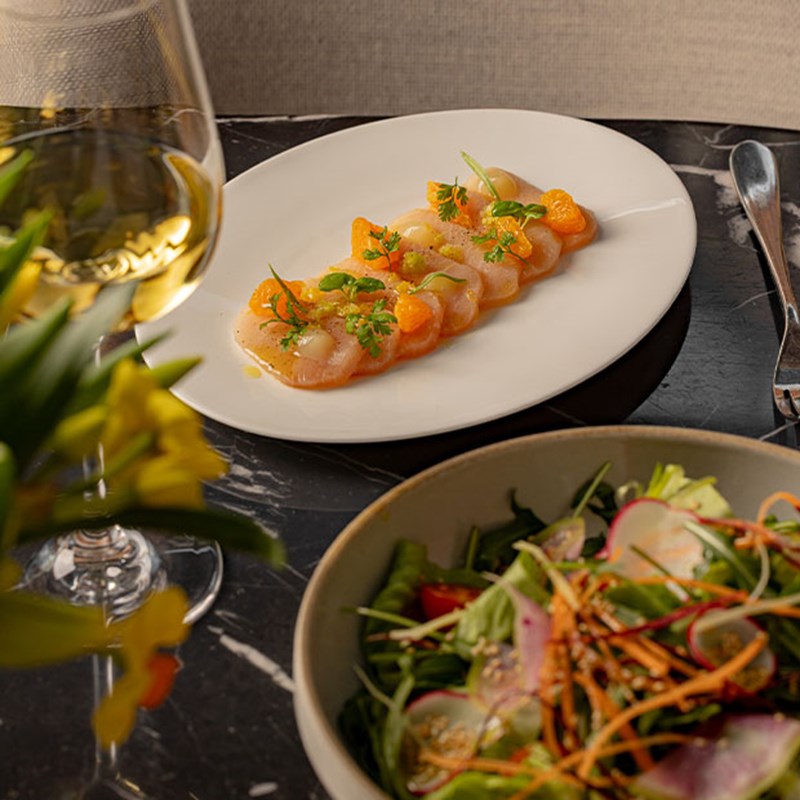 A refined meal of scallop carpaccio and fresh salad with a glass of white wine on a black marble table.