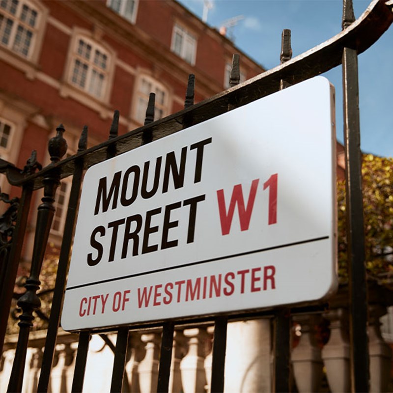 Close-up of Mount Street W1 road sign on wrought iron gate, with a red-brick Westminster building softly blurred in background.