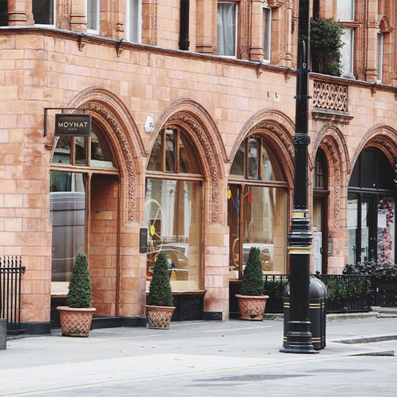 Luxury Moynat boutique with grand arched windows and terracotta brick facade, flanked by potted topiary trees on a quiet pavement.