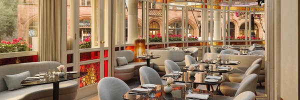 Light-filled dining area with curved seating, polished black tables, and panoramic windows looking onto red flowers and Mayfair’s historic buildings.