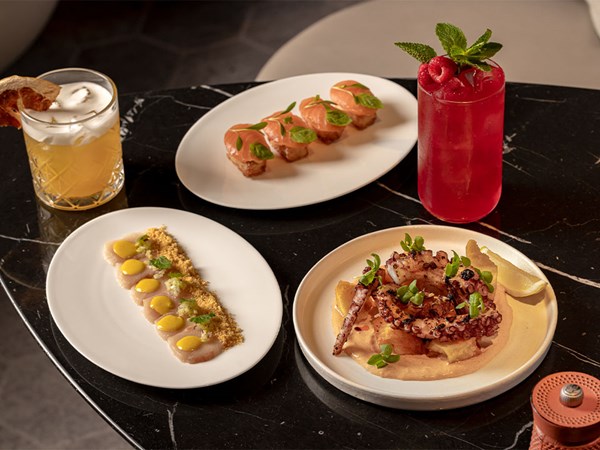 An elegant arrangement of modern small plates featuring grilled octopus, tuna tartare, scallop crudo, and two vibrant cocktails, served on a glossy black marble table.