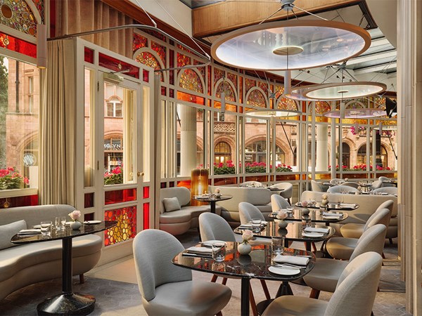 Stylish dining area at The Connaught with curved grey seating, black marble tables, and arched stained-glass windows framing views of historic Mayfair buildings.