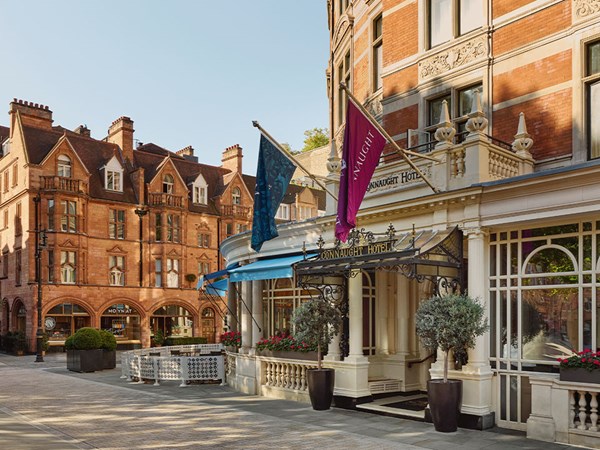 The grand entrance of The Connaught Hotel in Mayfair, featuring a wrought-iron canopy, flagpoles, and potted olive trees against a backdrop of red-brick architecture.
