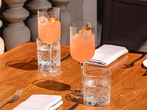Two tall stemmed glasses of a peach-pink cocktail with ice and floral garnish, served on a wooden outdoor dining table with crystal tumblers and folded napkins.