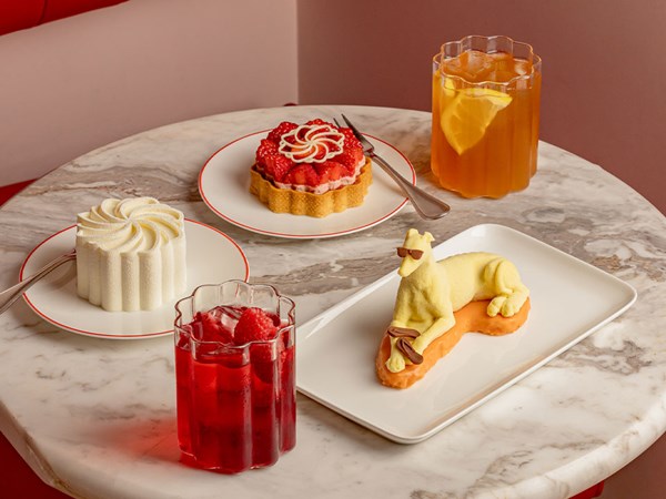 Marble table set with three playful summer pastries—including a white swirl mousse, a glazed strawberry tart, and a yellow dog-shaped dessert wearing sunglasses—served alongside a red berry drink and a citrus iced tea in faceted glasses.