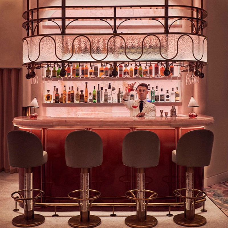 A view of the Red Room bar. 4 bar stools are tucked behind a pink onyx bar, with shelves of spirits lit up behind it.