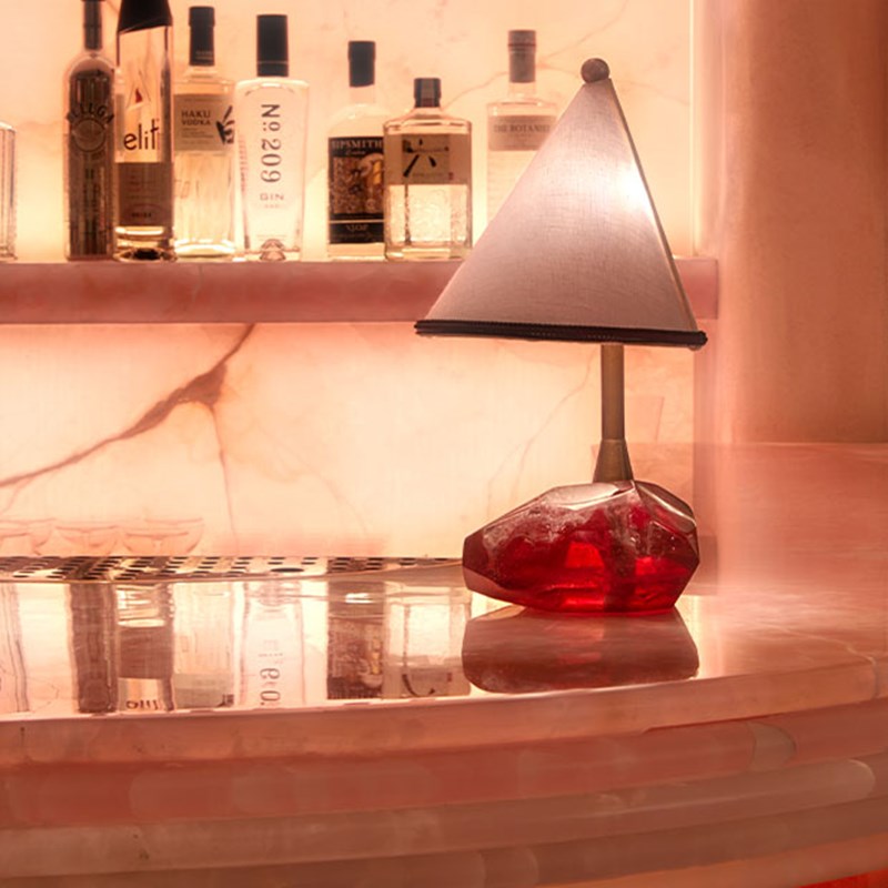 Triangular lamp with a red crystal-like base sat on the pink onxy bar, with spirits in the background.