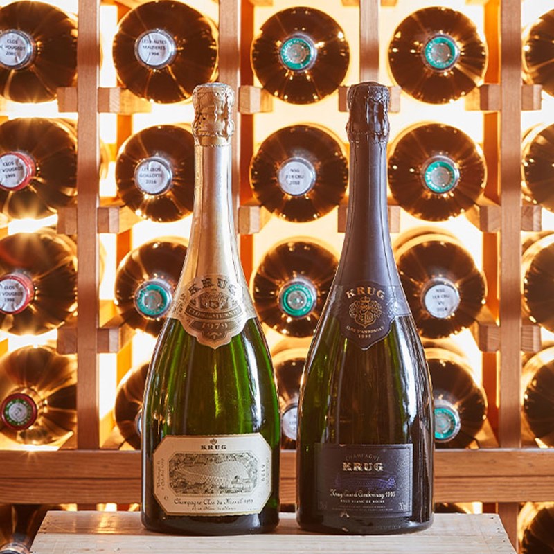 Two bottles of Krug champagne with bottles with a rack of red wine behind.