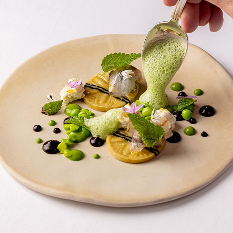 Helene Darroze dish with silver spoon pouring green jus onto a creation featuring peas, balsamic and cheese