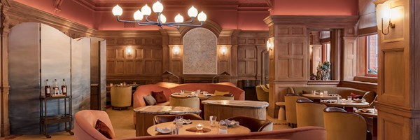 Spacious wood-panelled dining room with soft coral tones, ambient wall lighting, and curved velvet seating arranged beneath a central chandelier in a warmly lit setting.