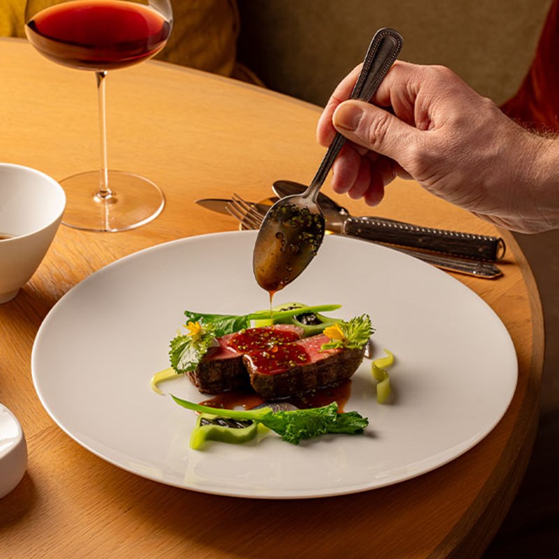Hand delicately drizzles sauce over seared meat garnished with vibrant greens and edible flowers, beside a glass of red wine on a round wooden table.