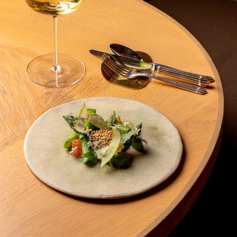 A fresh and artful dish featuring leafy greens, grains, and citrus on a stoneware plate, accompanied by white wine and silverware on a wooden table.