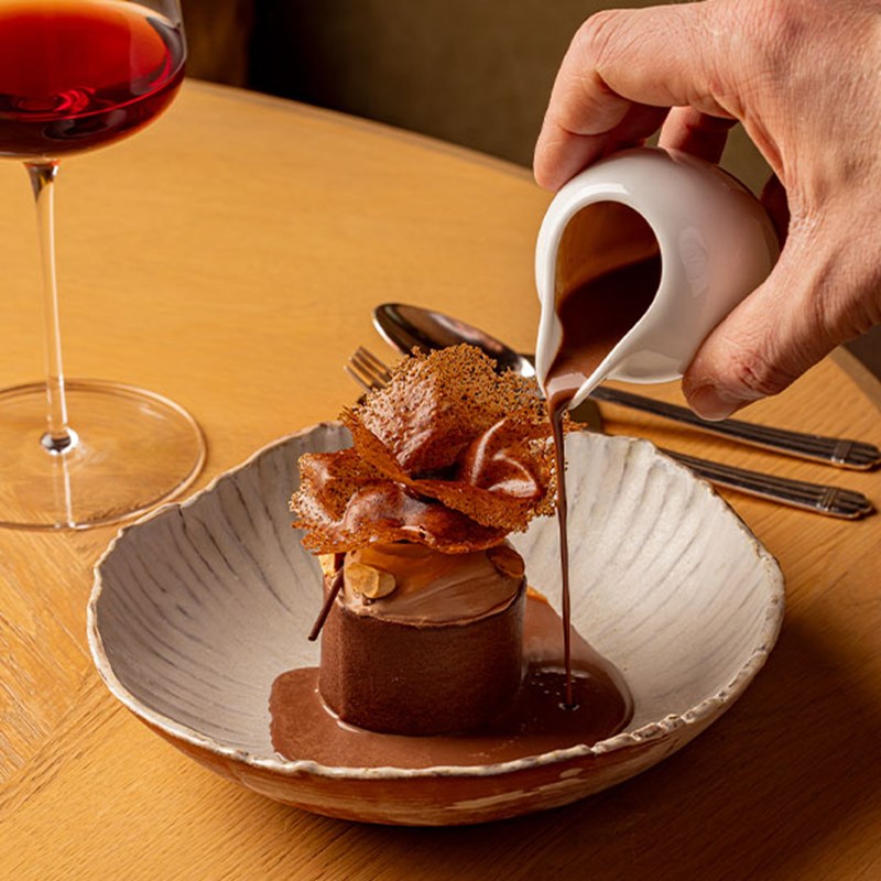 A hand pours rich chocolate sauce over an intricate plated dessert topped with delicate tuile, beside a glass of red wine on a wooden table.