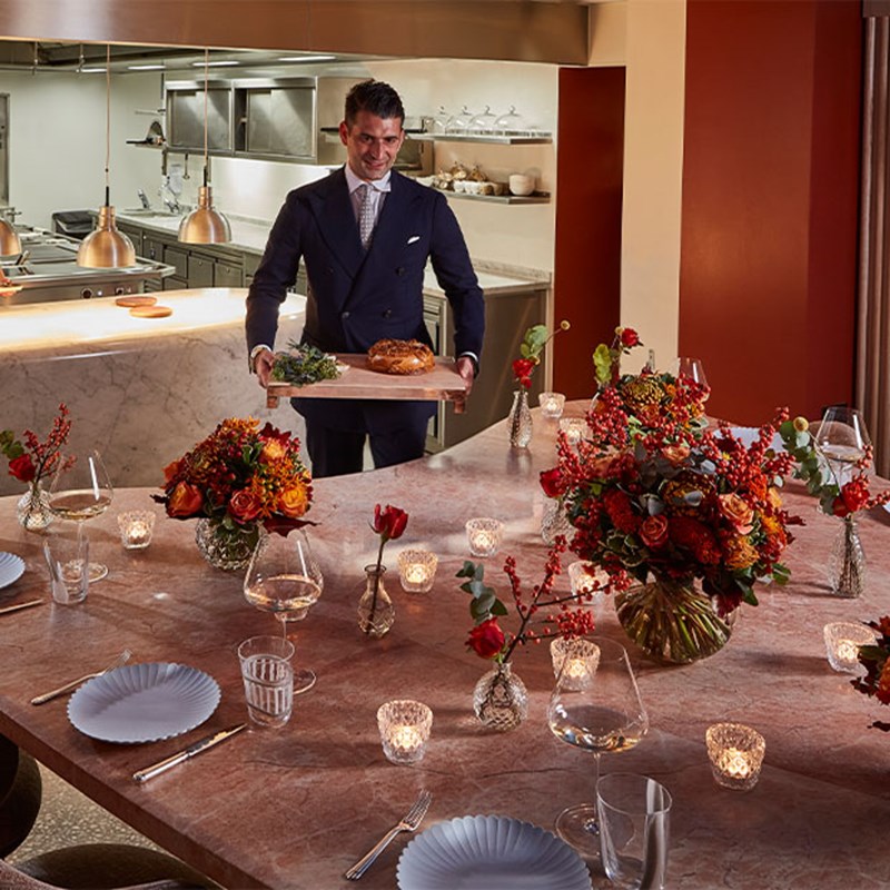 A smiling server in formal attire presents bread in a private dining room adorned with red flowers, candles, and marble counters near an open kitchen.