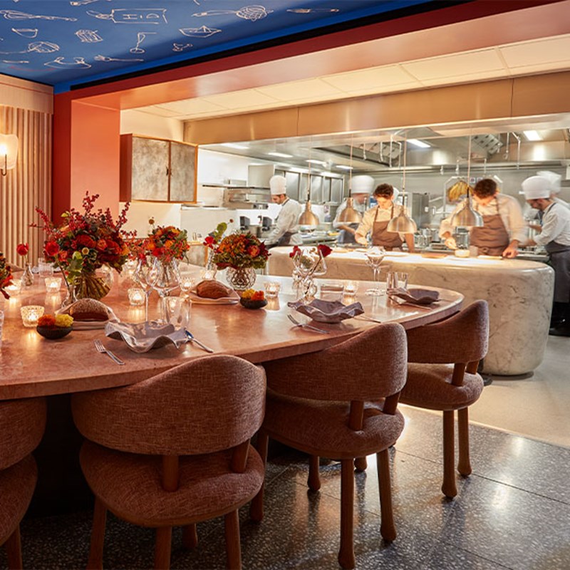 Private dining area with a pink marble table set with floral arrangements and candlelight, overlooking an open kitchen with chefs actively preparing dishes.
