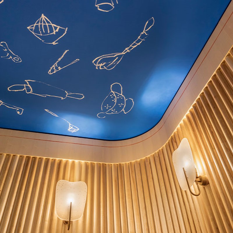 Blue ceiling with playful white line drawings of kitchen tools, surrounded by curved wooden panelled walls and softly glowing modern sconces.