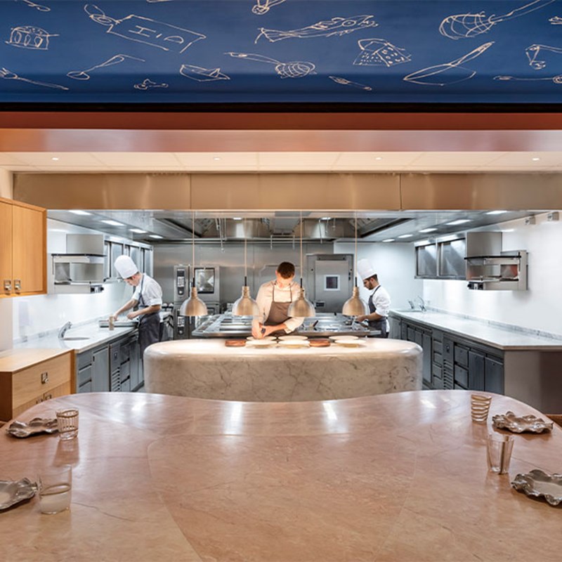 Open kitchen with chefs in uniform preparing food behind a marble counter, viewed from a round dining table beneath a ceiling illustrated with culinary motifs.