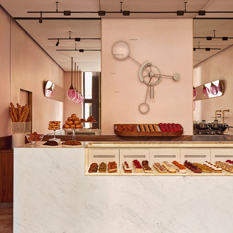 Elegant patisserie counter with white marble finishes, pink walls, and an array of pastries and breads displayed beneath a large, abstract clock installation.