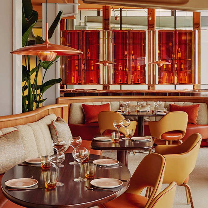 Contemporary restaurant interior with warm orange tones, curved seating, copper light fixtures, and amber glass panels behind round tables set for dining.