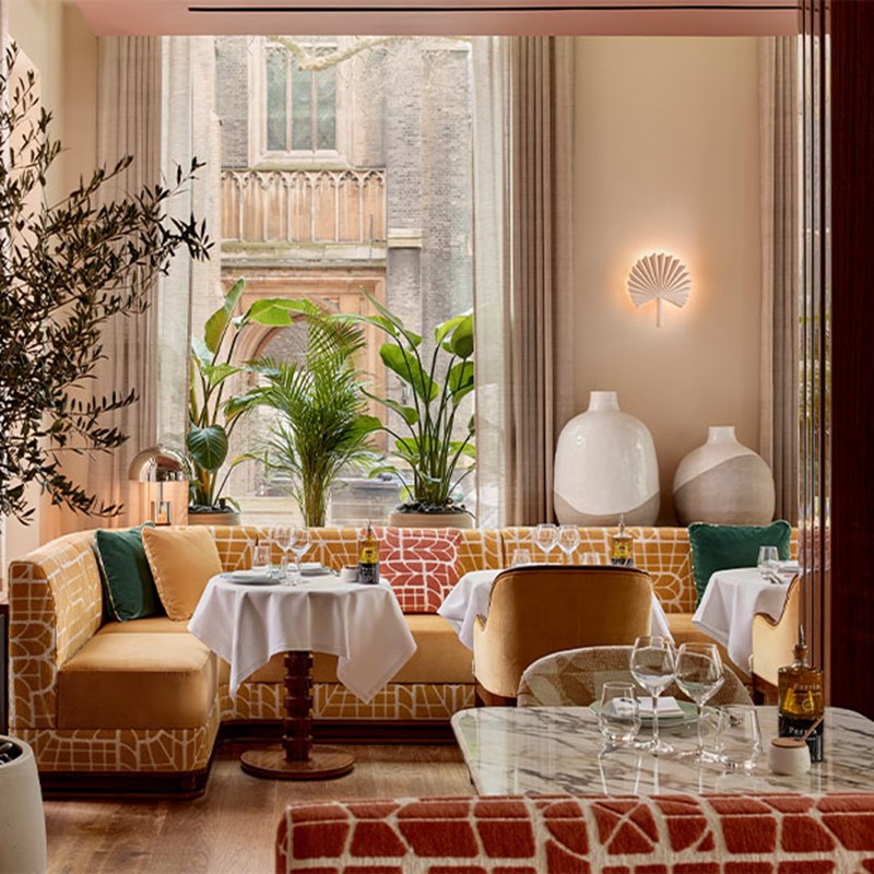 Sunlit dining alcove with patterned mustard sofas, potted greenery, and white tablecloths, offering a serene view of a historic stone church through tall windows.