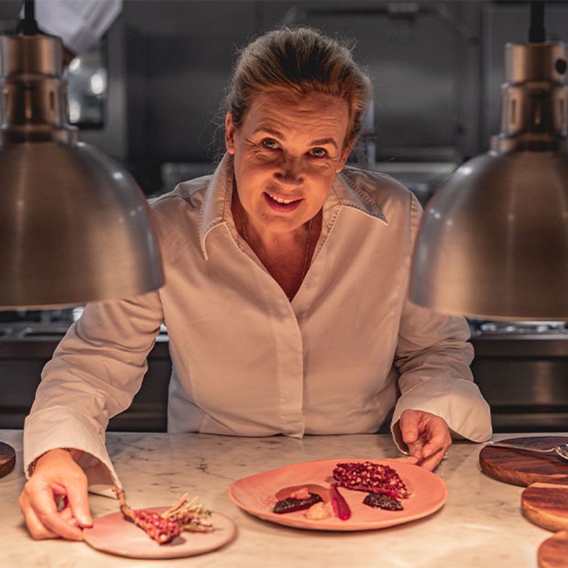 A female chef in a white jacket presents two artfully plated dishes under warm kitchen lighting, smiling as she leans over a marble counter.