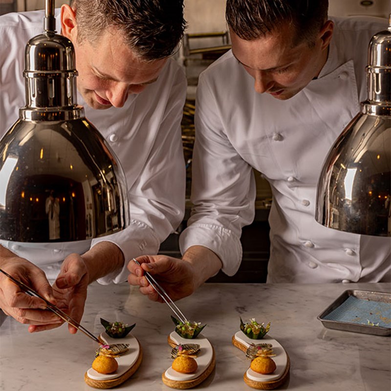 Two male chefs in white uniforms delicately use tweezers to garnish elegant hors d'oeuvres beneath stainless steel heat lamps in a professional kitchen setting.