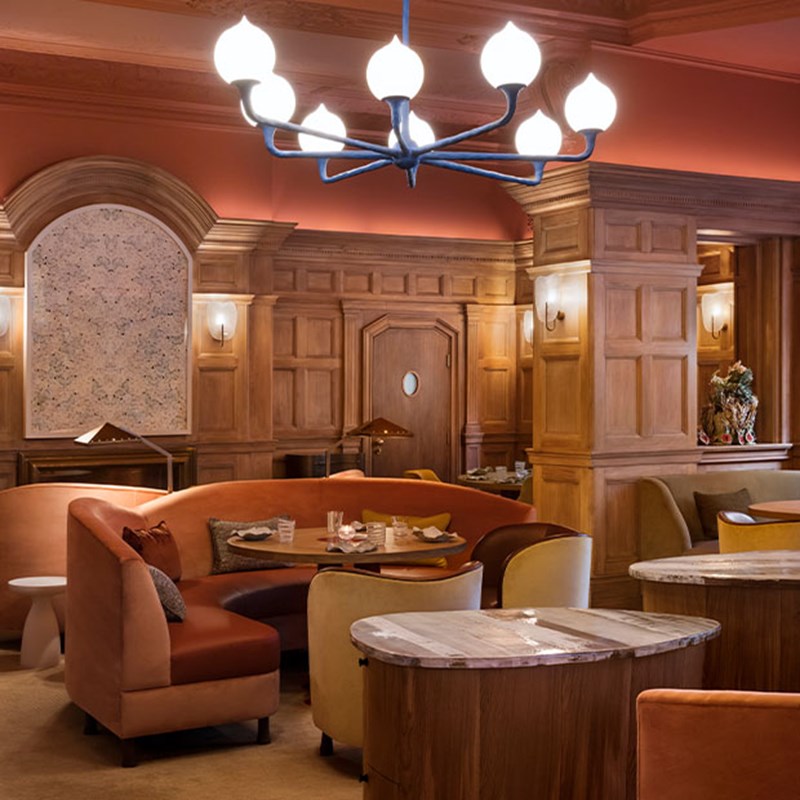 An inviting restaurant interior with peach-toned walls, plush seating, curved banquettes, and soft lighting beneath a contemporary chandelier and wood-panelled arches.