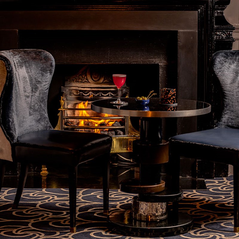 Two black velvet chairs surround a small round table beside a roaring fireplace, with a pink cocktail and snacks arranged in a refined setting.