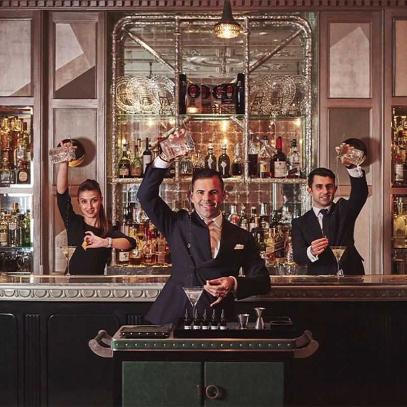 Three bartenders in formal attire pose enthusiastically behind a mirrored bar, mid-action as they shake, pour, and garnish stylish cocktails in glassware.