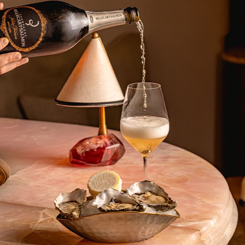 A glass of champagne being poured next to a lamp and bowl of fresh oysters on ice, atop a polished pink marble table in elegant lighting.