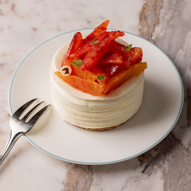 A plated dessert featuring a round white cake topped with vibrant slices of blood orange and delicate microgreens, placed next to a silver fork on a marble table.