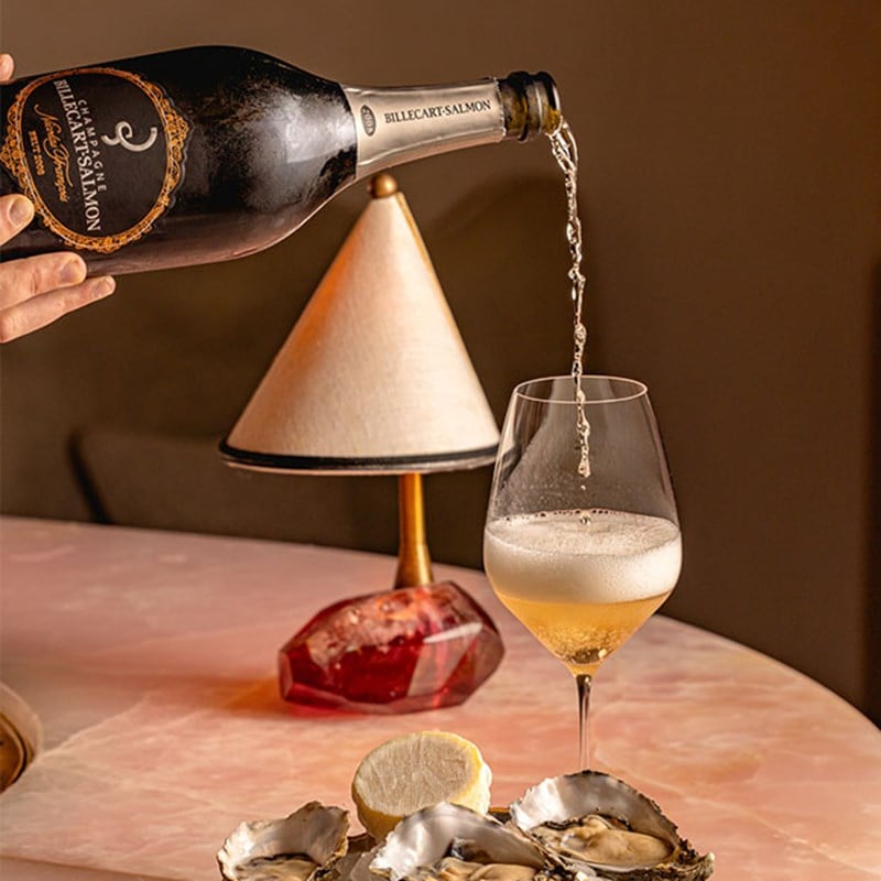 A hand pours Billecart-Salmon champagne into a tall glass on a marble table, with a plate of oysters, a halved lemon, and a small lamp in the background.