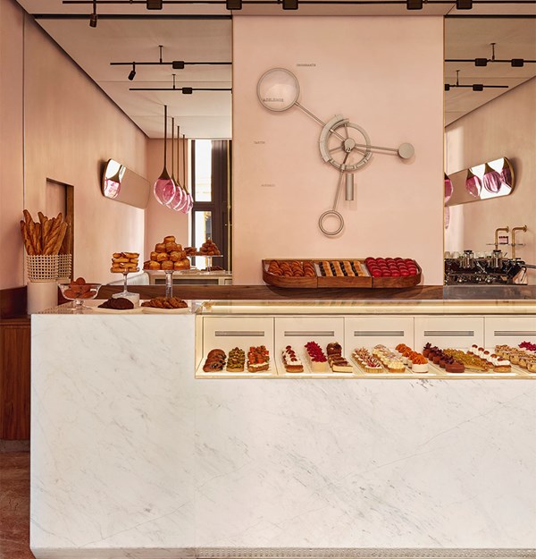 Elegant patisserie with blush pink walls, a marble counter displaying colourful pastries, and a sculptural wall clock behind fresh baked goods.