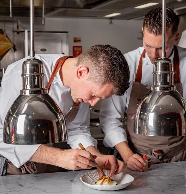 Two pastry chefs in white uniforms carefully plating a dessert under heat lamps in a professional stainless-steel kitchen environment.