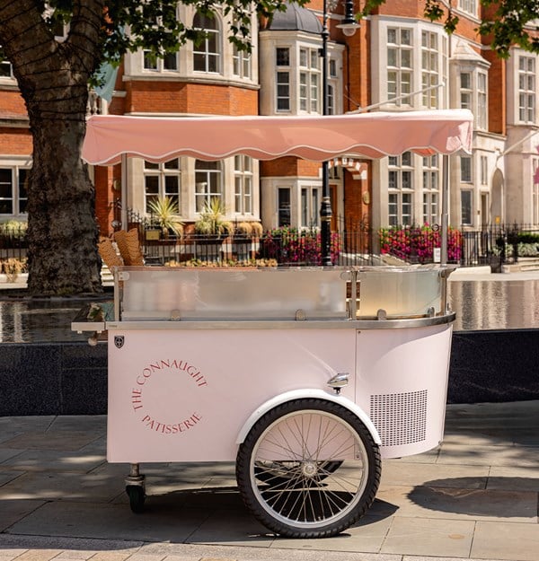 A pastel pink Connaught Patisserie cart with waffle cones parked on a sunny pavement, with elegant red-brick buildings in background.