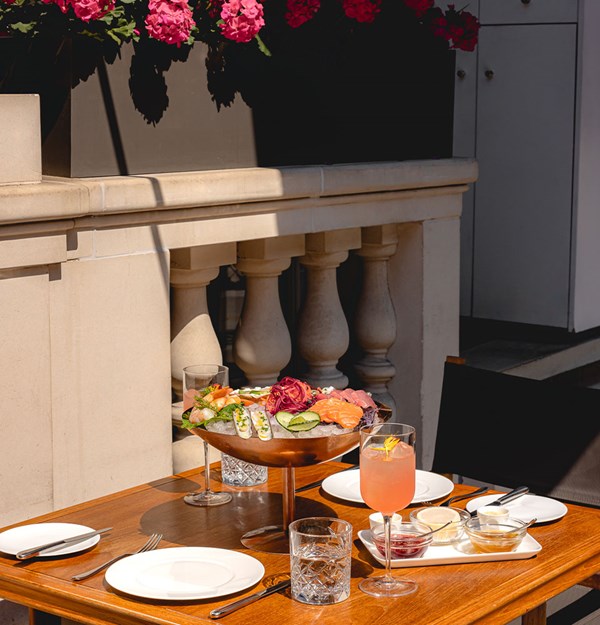 Sunlit wooden table on a terrace set with plates, cutlery, drinks, and a large seafood platter beneath blooming pink flowers.