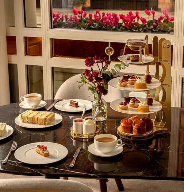 Afternoon tea setting with tiered trays of pastries, scones, and sandwiches, accompanied by tea and coffee on a marble table beside a window of pink flowers.