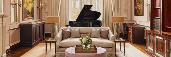 Elegant living room with a cream sofa, pastel cushions, round ottoman with flowers, and a grand piano by tall curtained windows, surrounded by framed art and dark wood furniture.