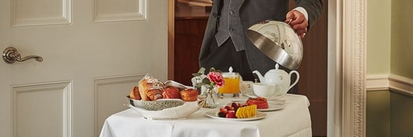Hotel server in a suit lifting a silver cloche to reveal a breakfast table with pastries, fruit, smoked salmon, juice, tea set, and a small vase with a pink flower.