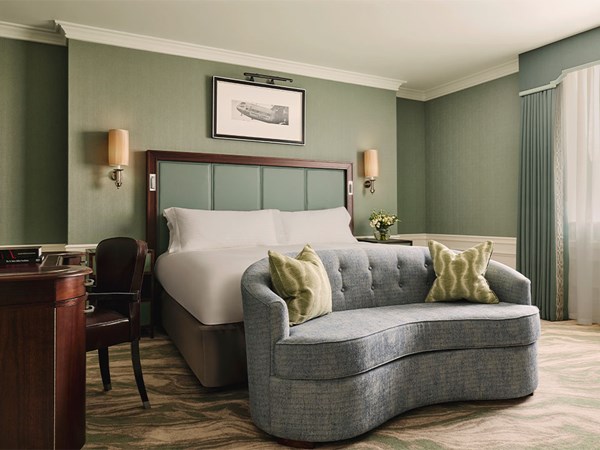 Serene bedroom with king bed, soft green walls, curved loveseat at the foot, and a writing desk.