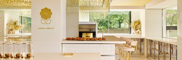 A bright, elegant patisserie with gold accents, glass-domed pastries, and a central counter overlooking a leafy garden through large windows