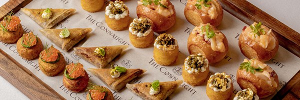 Assorted gourmet canapés including salmon roe bites, samosas, caviar-topped rounds with gold leaf, and brioche rolls with seafood filling, arranged on a wooden tray lined with The Connaught paper.