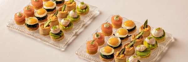 Selection of canapés including smoked salmon rolls, mini fried quail eggs on spinach, asparagus tartlets, and avocado with poached egg, neatly arranged on glass trays.