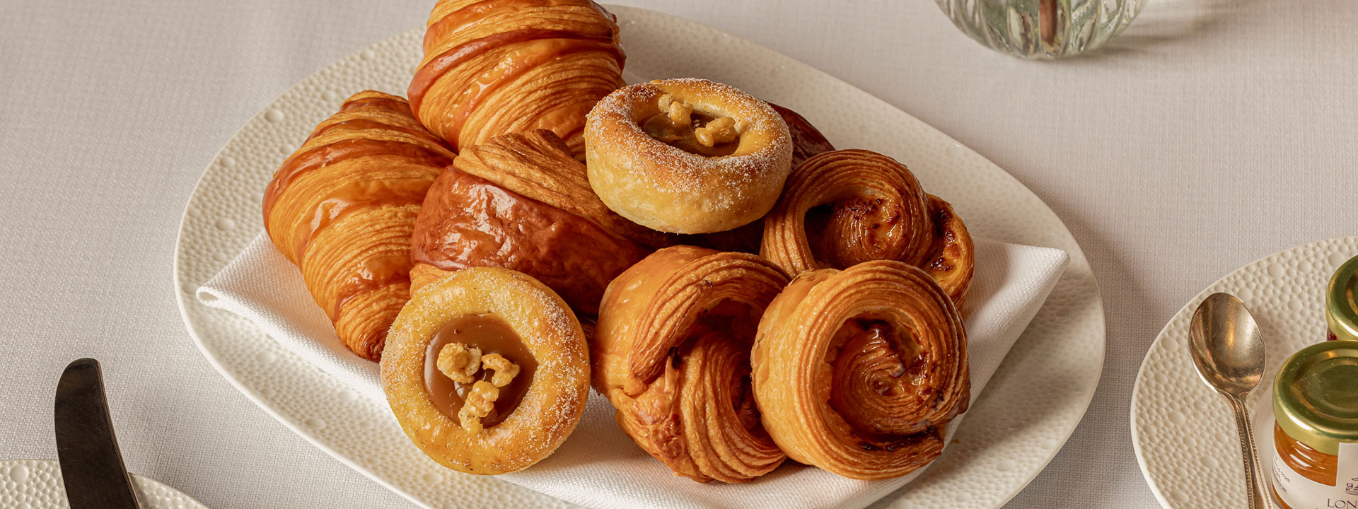 Plate of assorted breakfast pastries including croissants, swirls, and caramel walnut danishes, served on a white textured dish with a linen napkin.