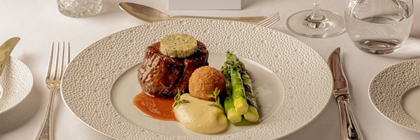 Beef fillet topped with herb butter, served with asparagus, potato purée, a croquette, and a rich jus on a white textured plate.