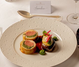 Two slices of lamb loin wrapped in pastry with herb filling, served with stuffed aubergine, green purée, and a glossy jus.