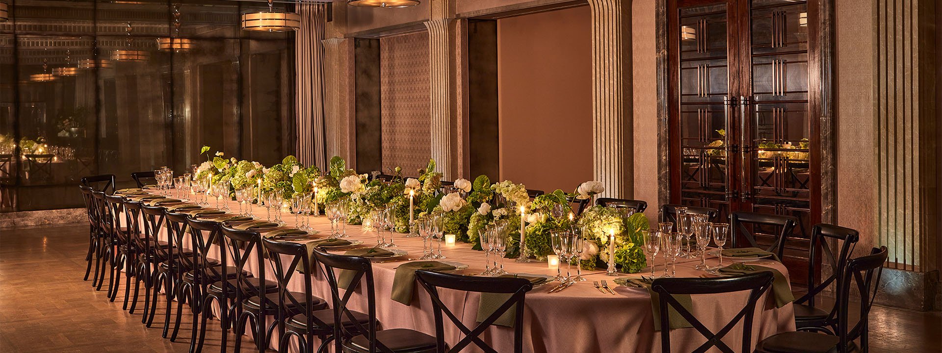 A luxurious dining room is elegantly set for an event, featuring round tables with white tablecloths, floral centerpieces, and candlelight, surrounded by stylish chairs with wicker backs.