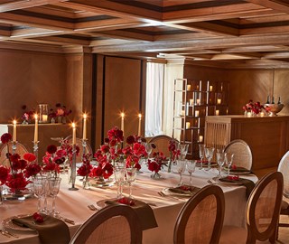 A long, elegantly set dining table with floral arrangements and candles is surrounded by dark wooden chairs in a sophisticated room with wooden floors and ambient lighting.